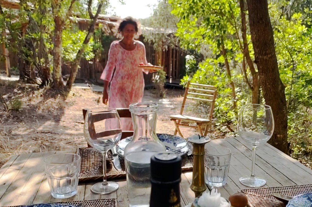 Woman in a pink floral dress carrying a plate walking towards an outdoor wooden table set with glasses and a water carafe in a shaded garden area.