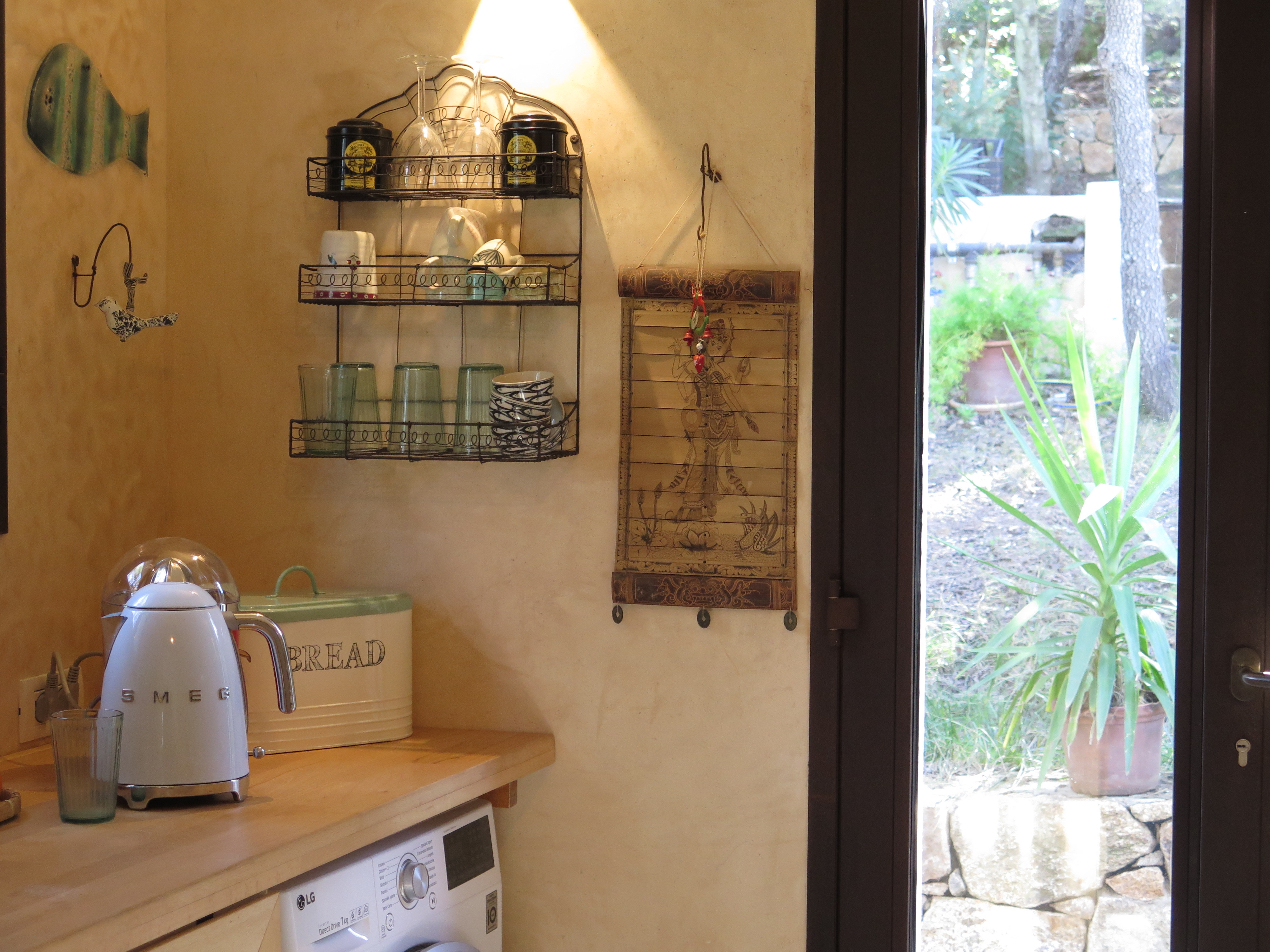 Kitchen corner with a white SMEG kettle, a green bread box, wall-mounted shelves holding glasses and cups, a hanging decorative scroll, and a door opening to a garden with potted plants.