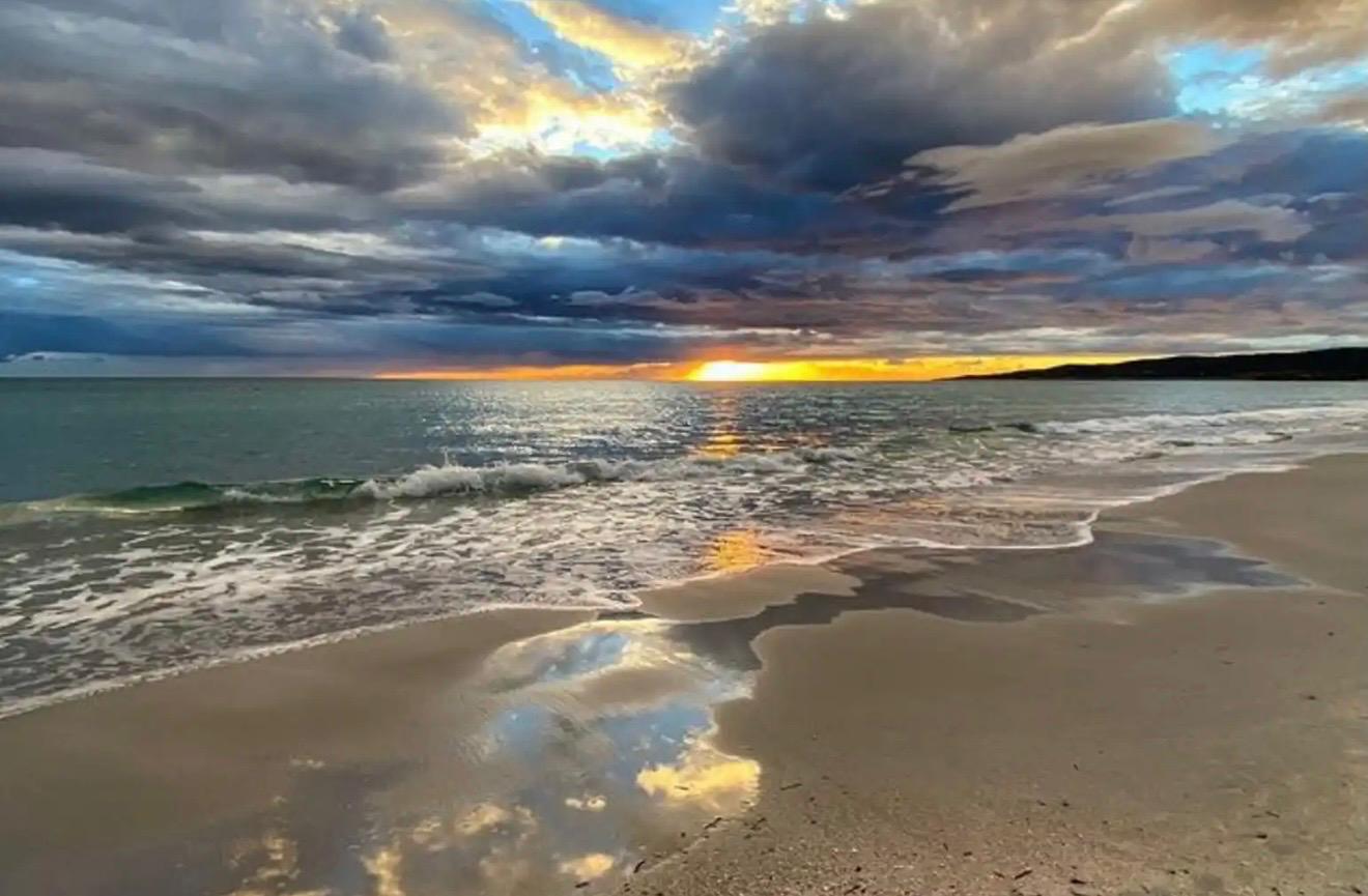 Ocean beach at sunset with waves gently rolling onto the sandy shore under a dramatic cloudy sky.