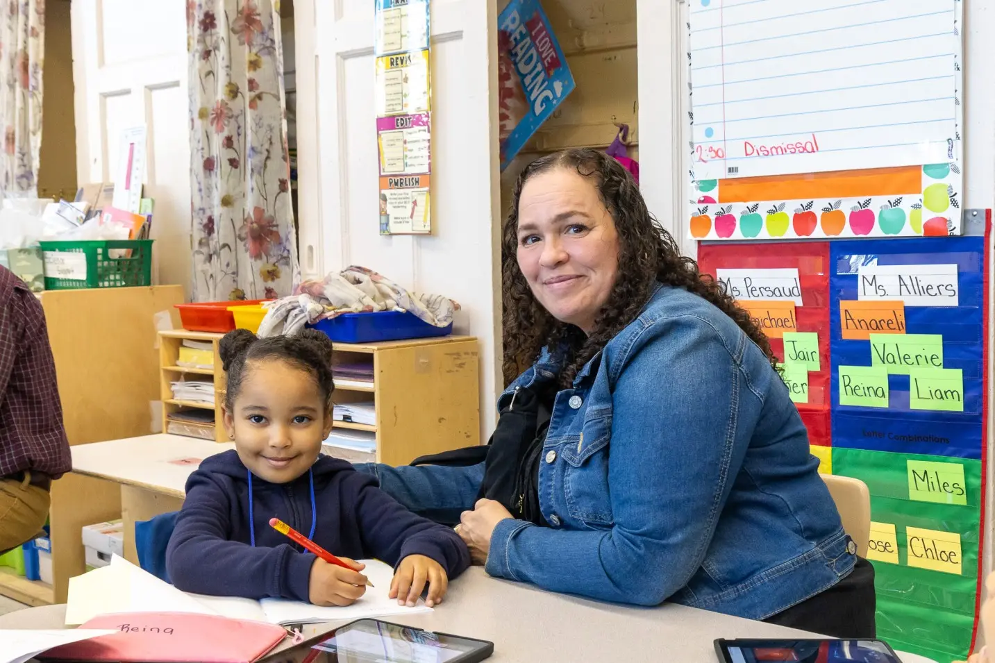 Teacher and young student smiling together at classroom desk writing