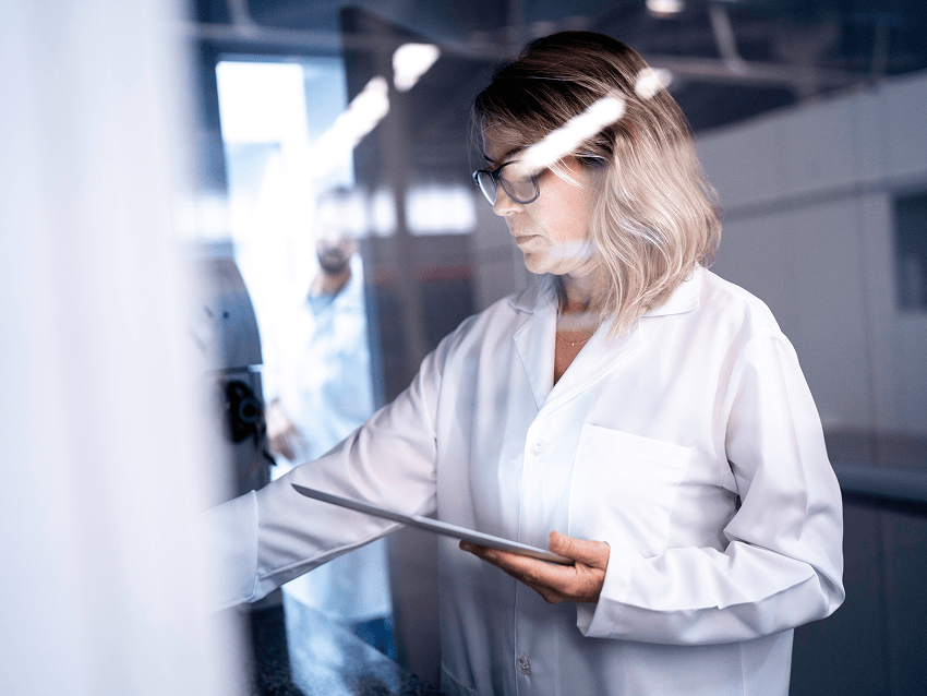 Image of a woman in a lab coat holding a tablet, looking at a piece of equipment in a lab.