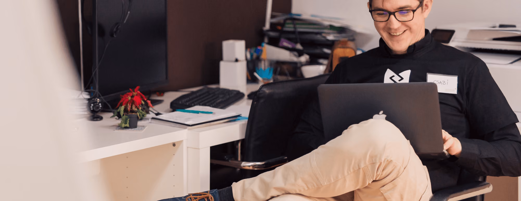 Man wearing glasses sitting at a desk, smiling while using a laptop in an office setting.