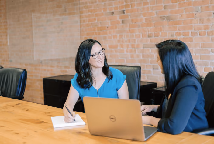 Two women having a meeting at a wooden table with a laptop and notepad in a brick-walled room.