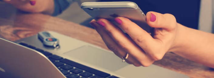 Person with pink nail polish holding smartphone over a laptop keyboard on wooden desk.
