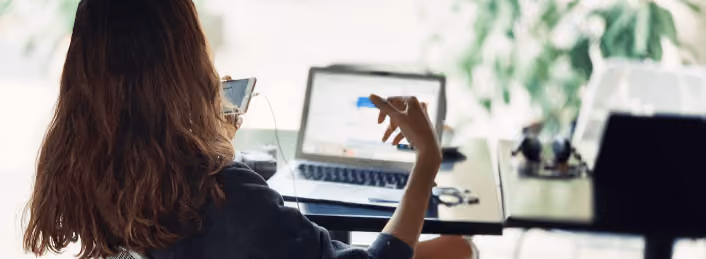 Person with long hair using smartphone and laptop at a glass desk with headphones on the side.