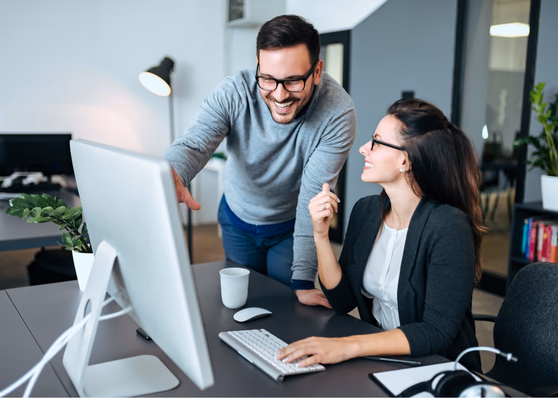 Man and woman wearing glasses smiling and discussing something on a computer screen in an office.
