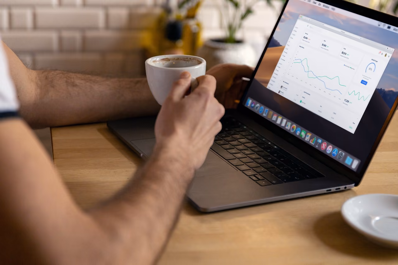 Person holding a white cup of coffee near a laptop showing a data analytics dashboard on screen.