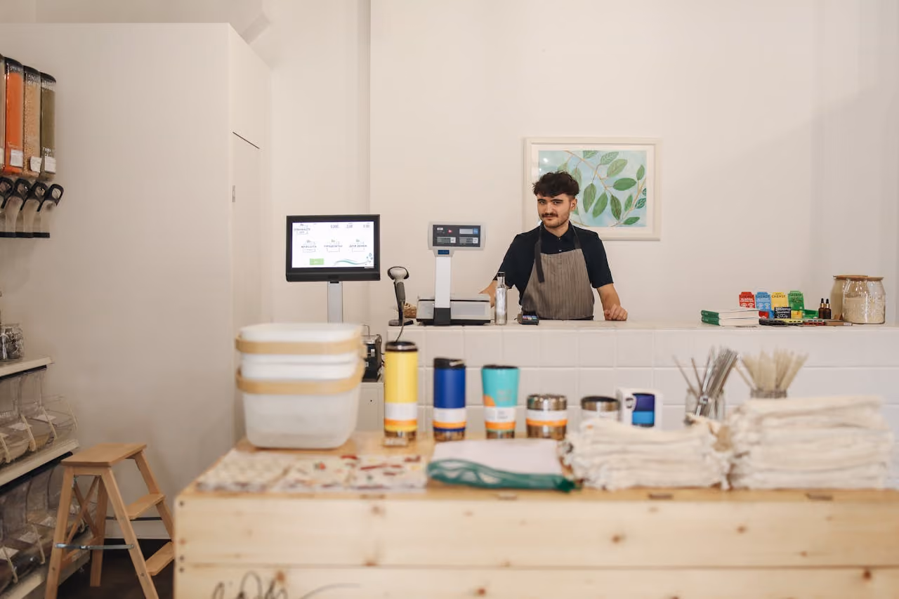 Young man in apron behind counter in store with reusable containers and eco-friendly products on display.