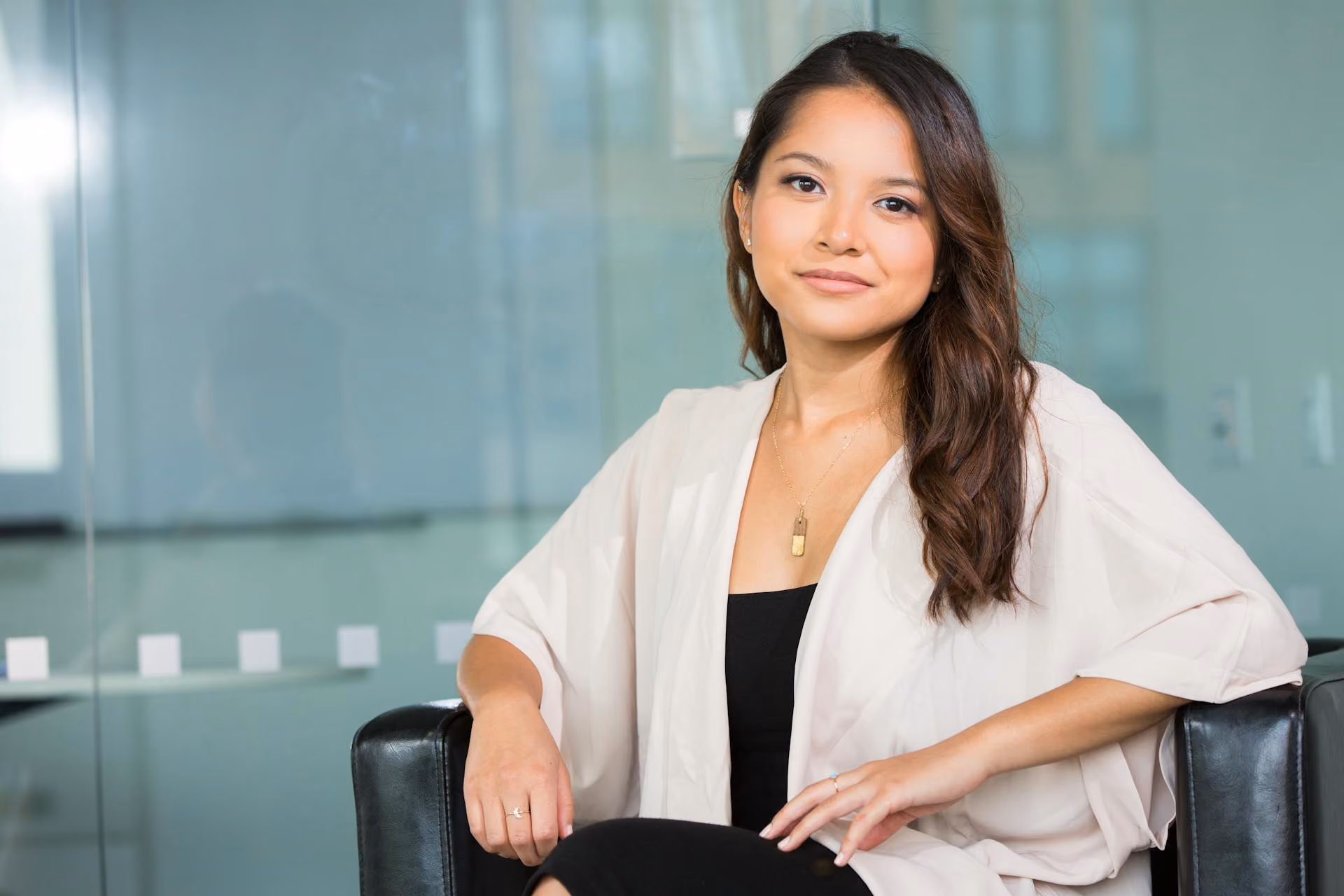 Woman with brown hair sits confidently on black chair wearing white cardigan and black dress.