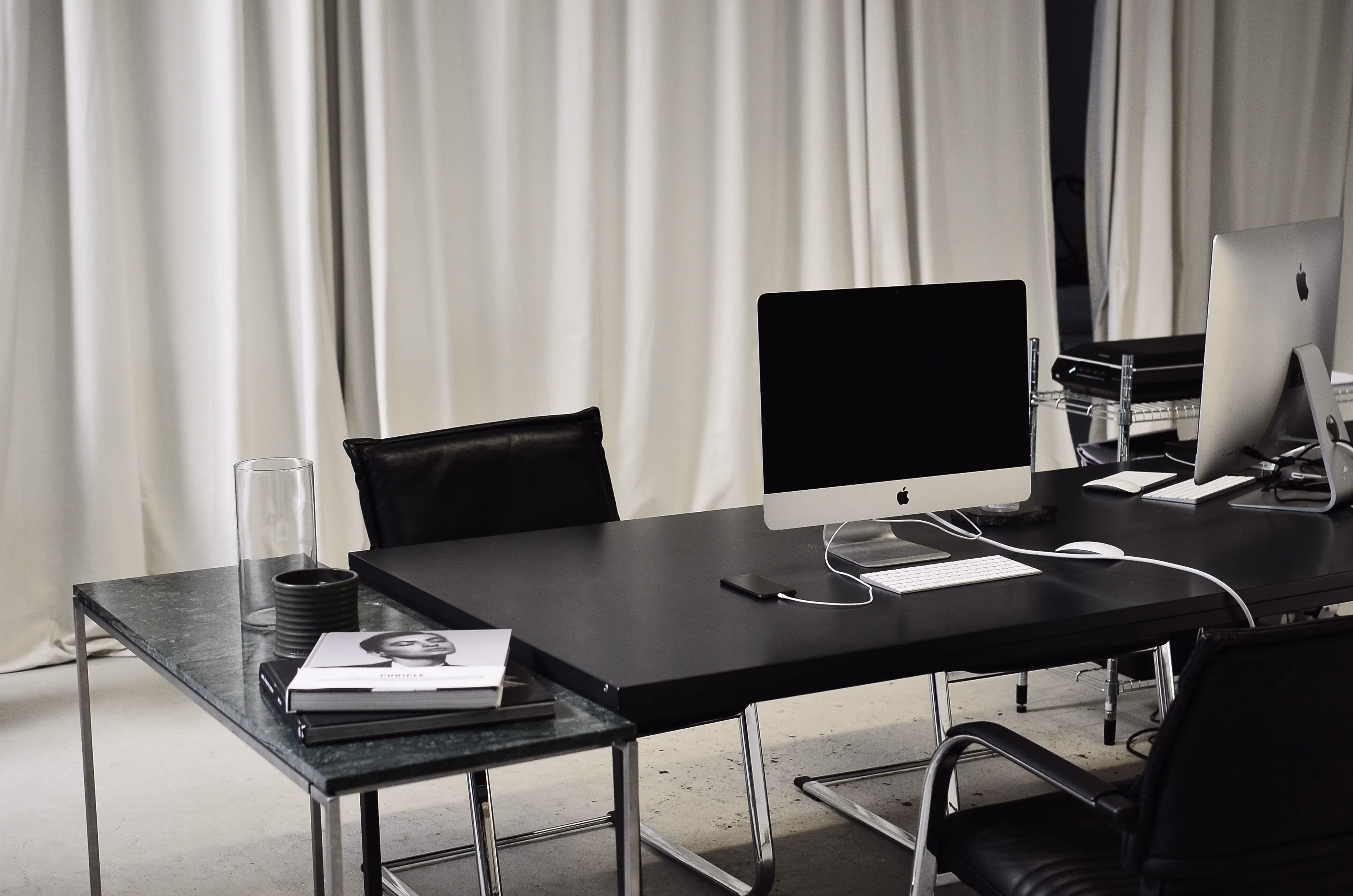 Modern office desks with iMac computers, black chairs, books, and a glass cup in front of white curtains.