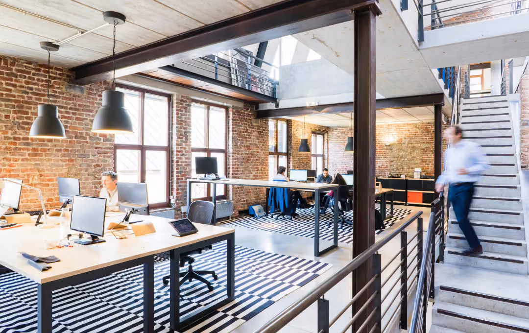 Modern open office with brick walls, large windows, desks with computers, and a person walking down stairs.