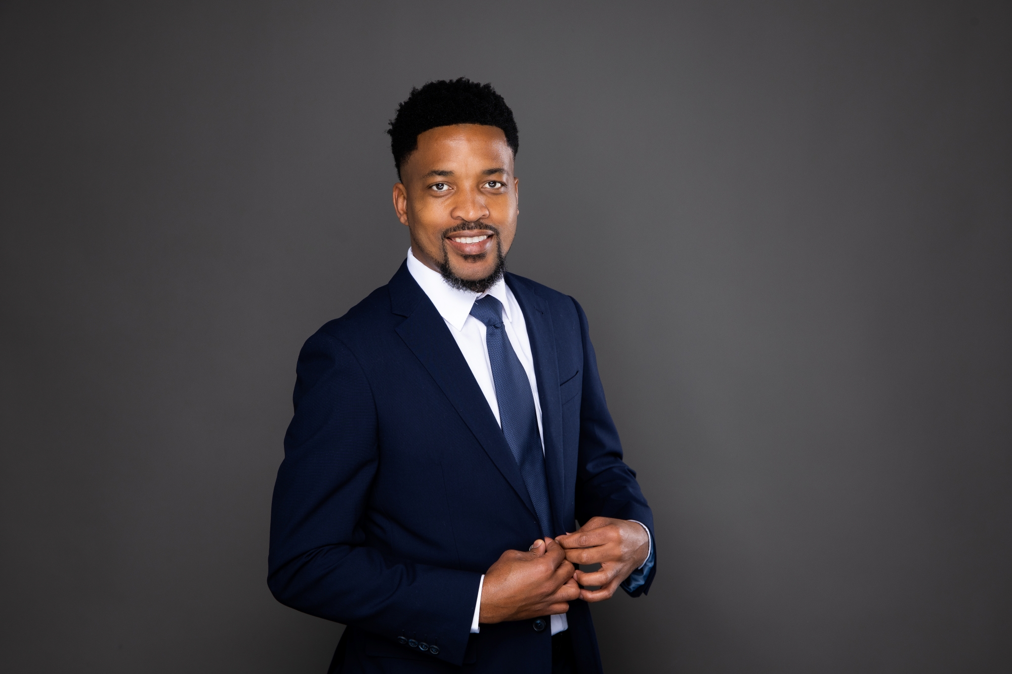 Smiling man in a navy blue suit and tie adjusting his jacket against a grey background.
