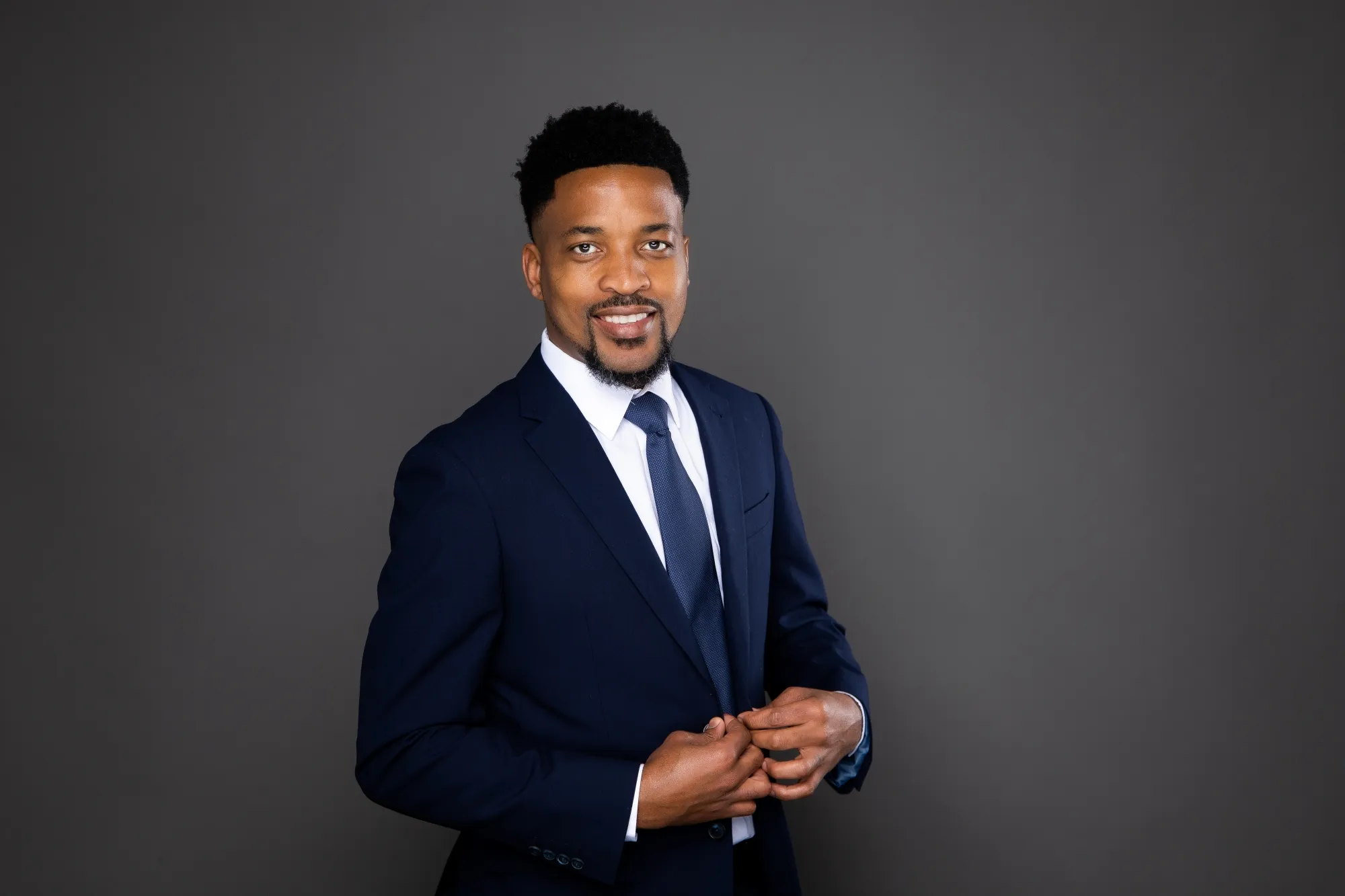 Smiling man in a navy blue suit and tie adjusting his jacket against a grey background.
