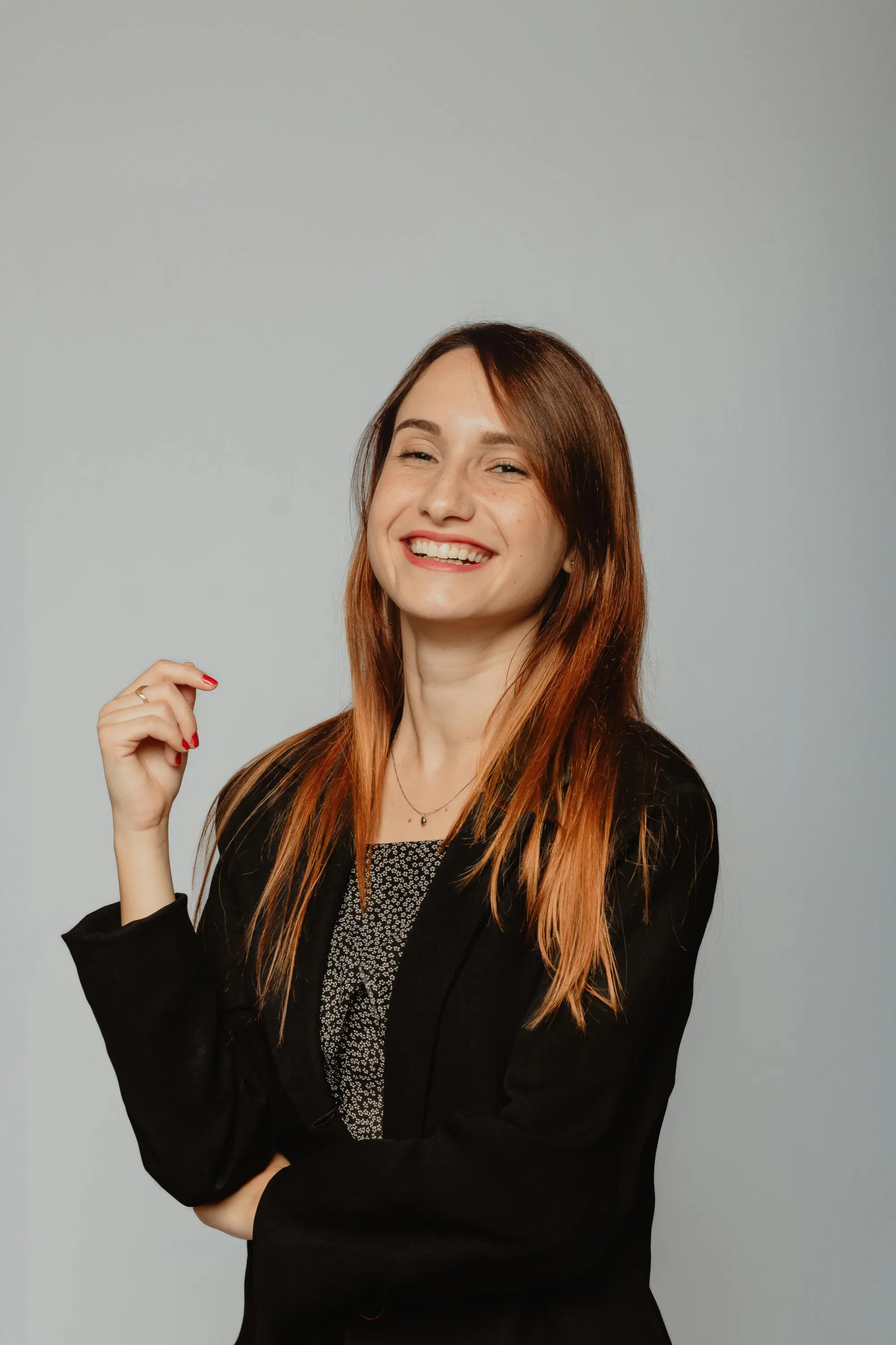 Smiling woman with long light brown hair wearing a black blazer and patterned top against a plain grey background.