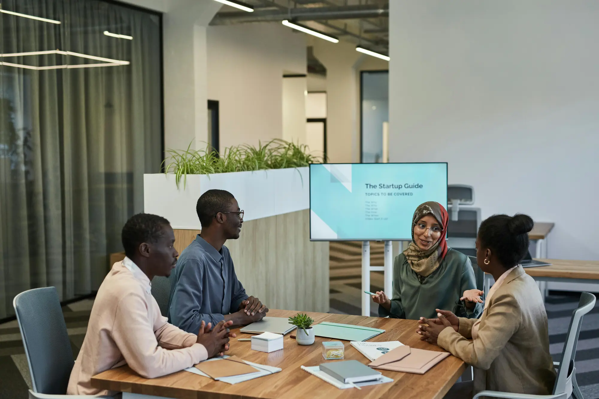 Four diverse professionals seated around a wooden table engaged in a discussion with a screen displaying 'The Startup Guide' in the background.