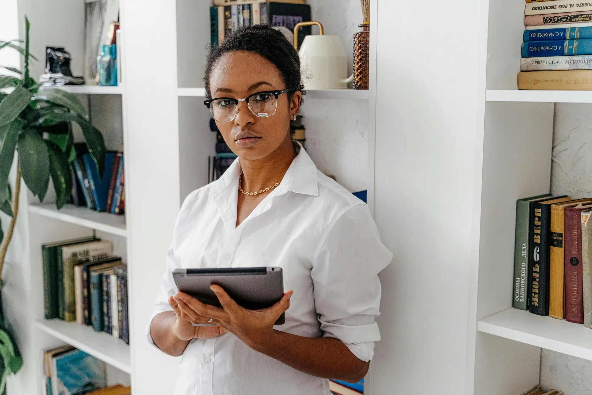Woman with glasses and white shirt holding a tablet, standing in front of a bookshelf filled with books.