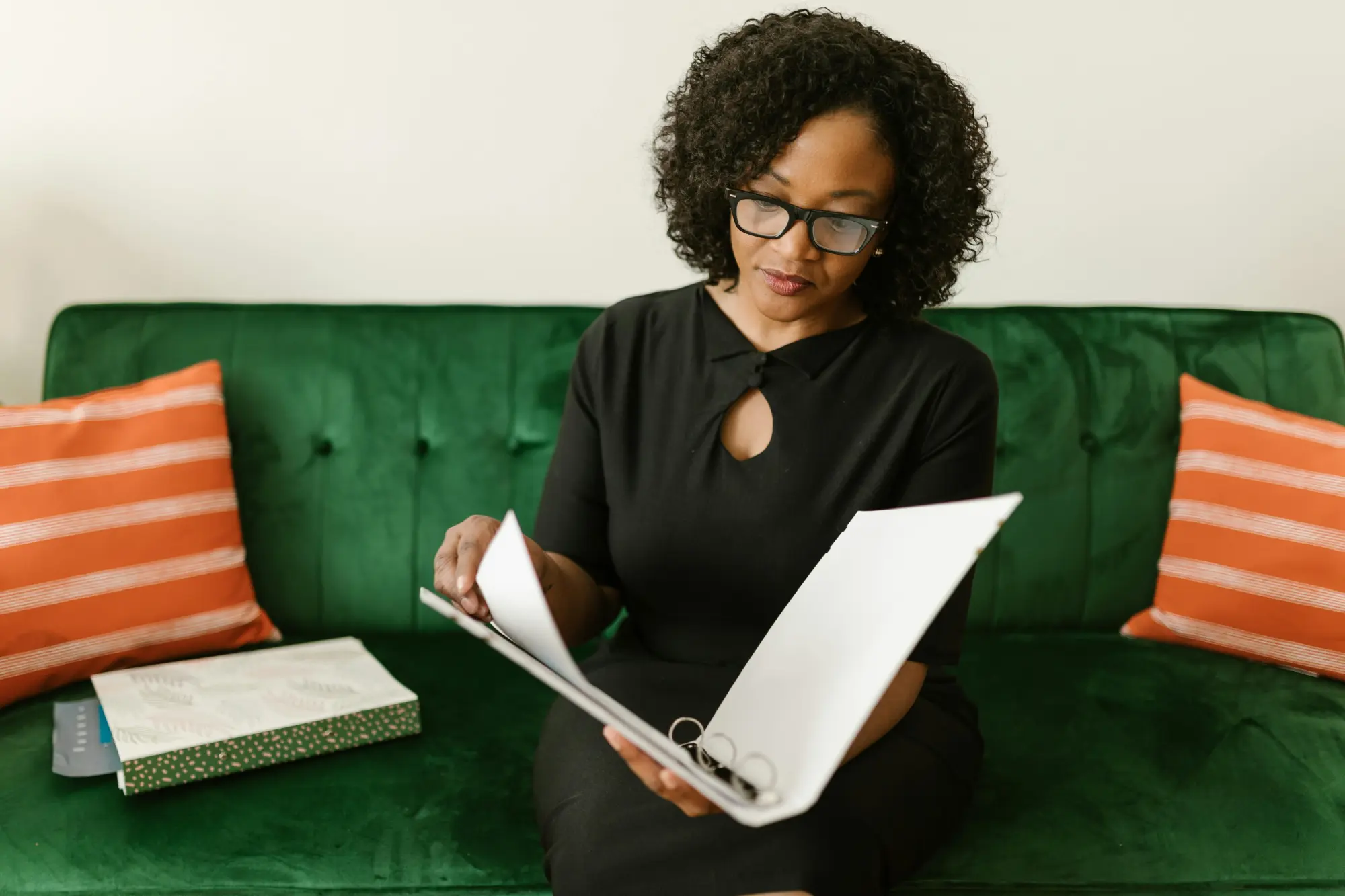 Woman with glasses sitting on a green couch and reviewing papers in a binder.