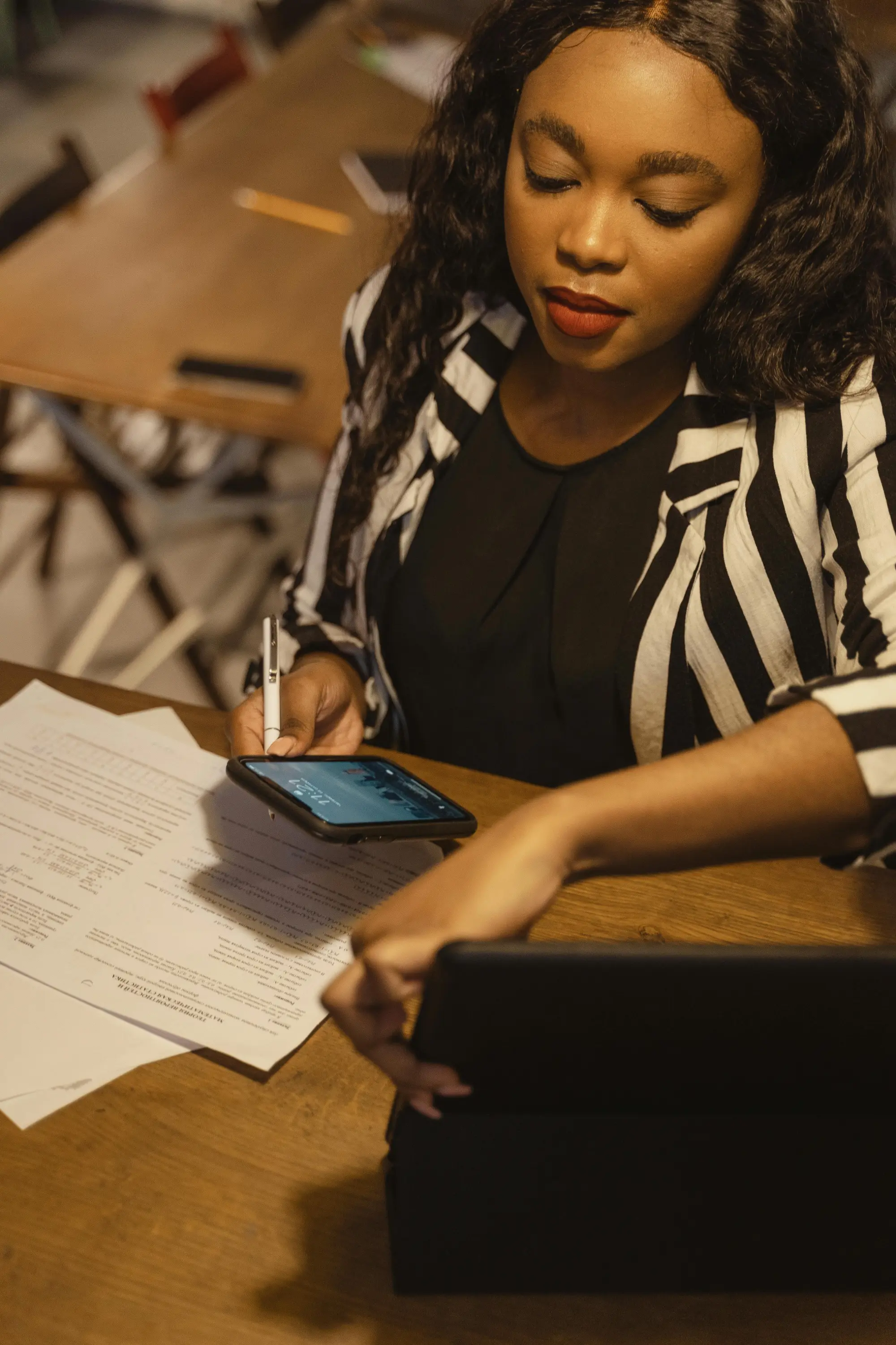 Woman in a black and white striped jacket working at a wooden desk with papers, holding a phone and a pen, looking at a tablet.