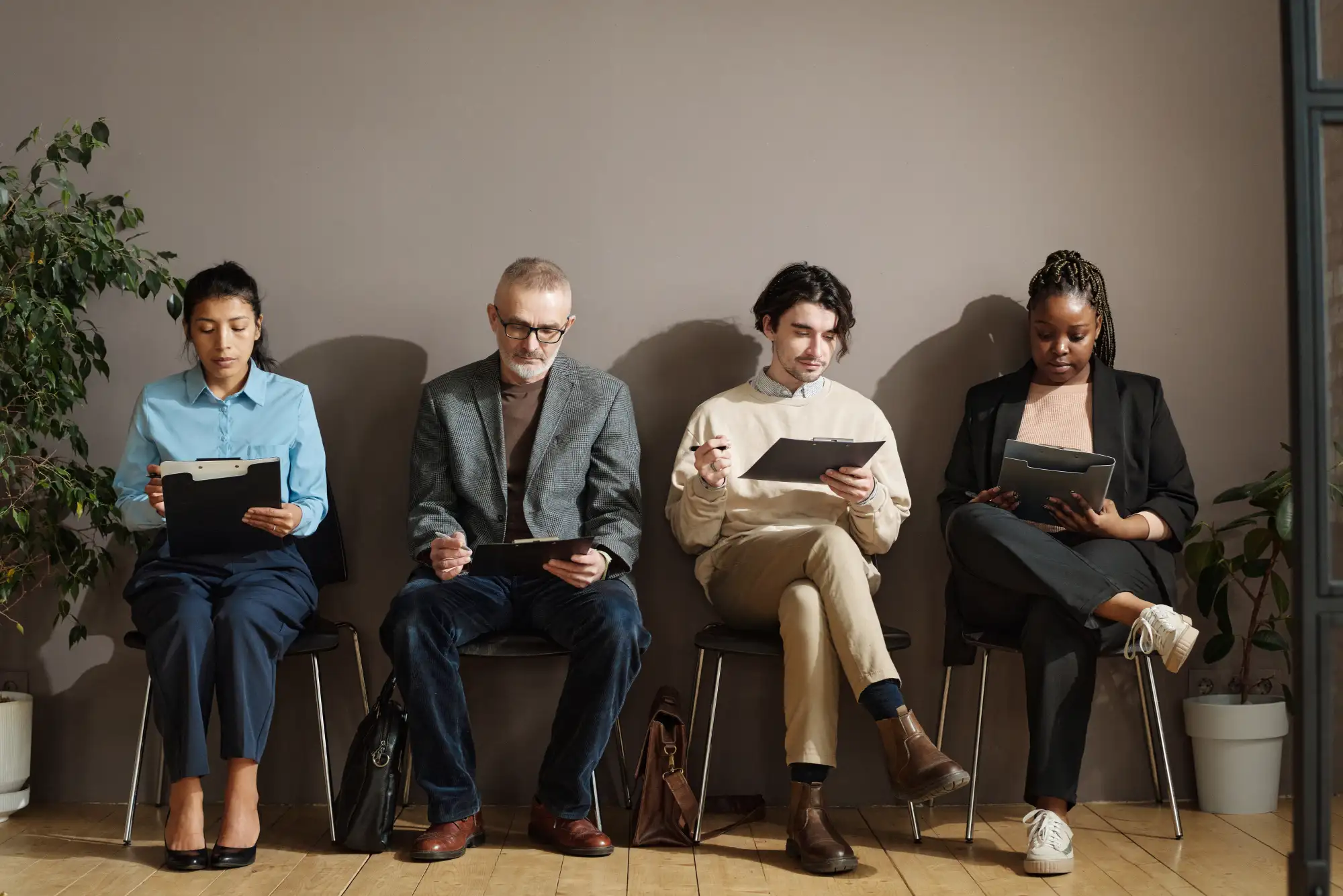 Four diverse people sitting on chairs against a wall, filling out forms on clipboards, waiting.