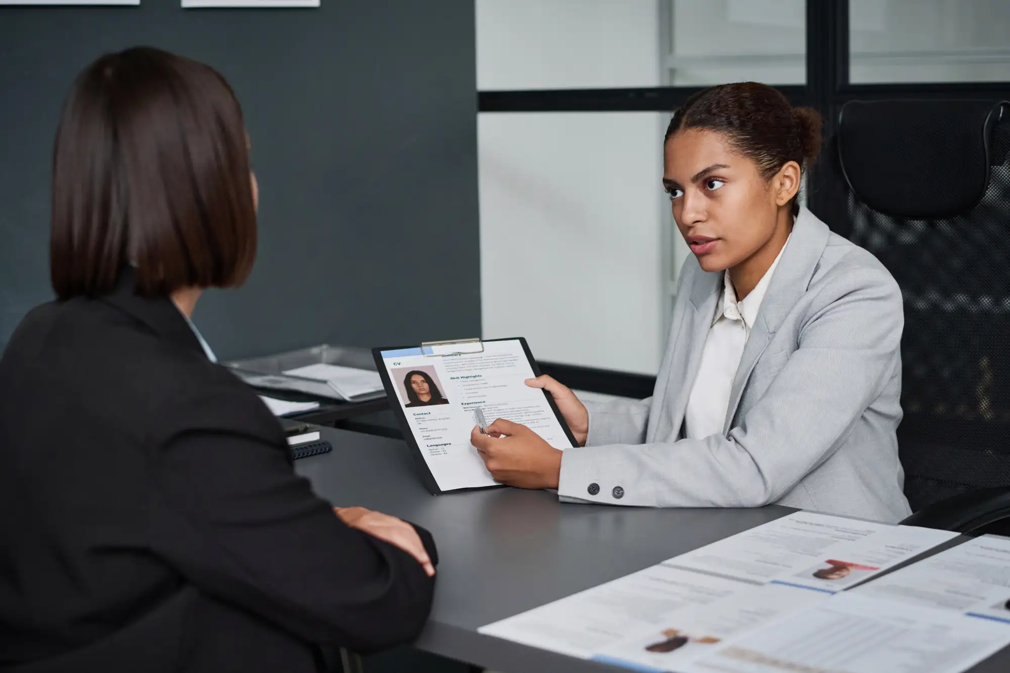 Woman in grey blazer conducting a job interview, pointing at a CV with a pen while talking to a candidate at a desk.