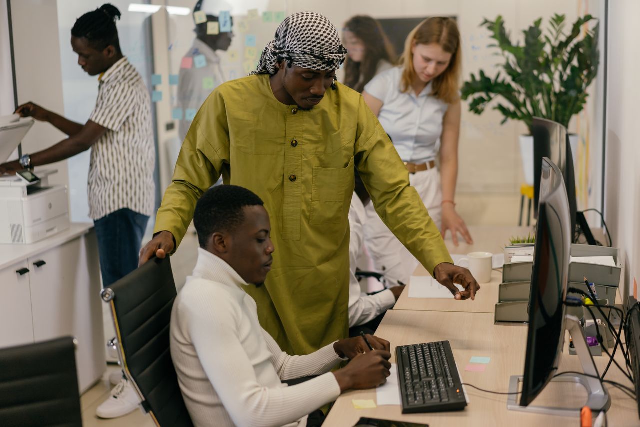 A supervisor leans over to guide a team member at his computer workstation while colleagues work in the background.