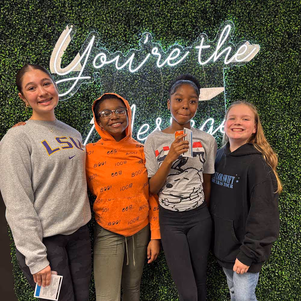 Four smiling teenage girls standing in front of a green hedge wall with neon sign reading 'You're the blessing', holding cards or tickets.