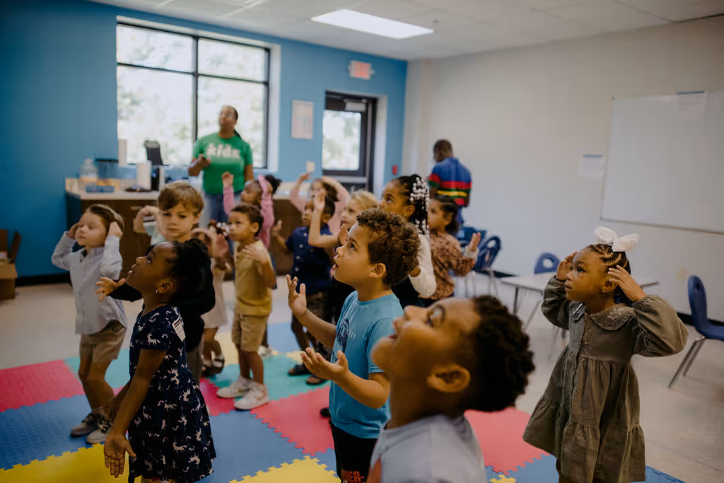 Group of young children standing and raising their hands in a colorful classroom with teachers in the background.