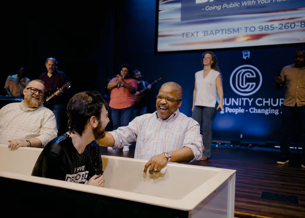 A man smiling and placing his hand on another man's shoulder during a baptism ceremony in a church with a band and people clapping in the background.