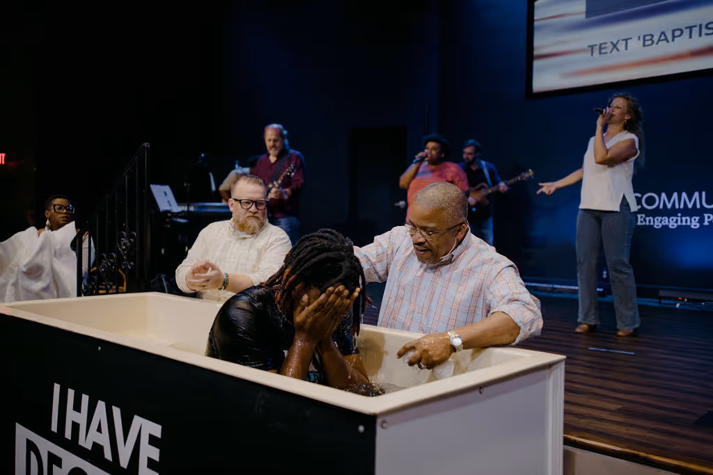 Person being baptized in a white baptismal pool inside a church with a man assisting and musicians and a singer performing on stage in the background.