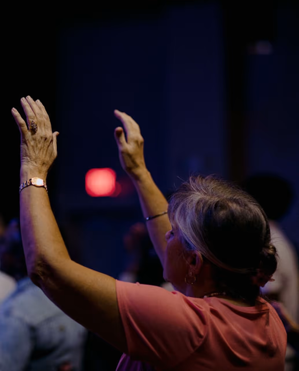 Woman with blonde hair in a pink shirt raising her hands in a dimly lit indoor setting.