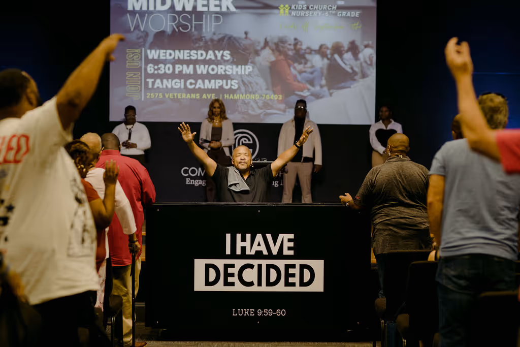 Man with raised hands behind a podium that reads 'I HAVE DECIDED' in a worship service with attendees raising hands.