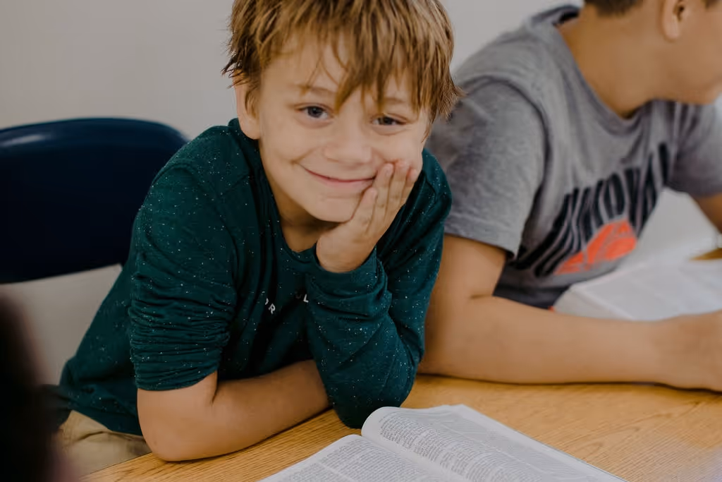 Smiling boy resting his chin on his hand sitting at a table with an open book.