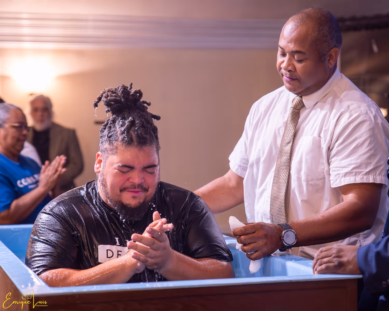 Man with dreadlocks being baptized in a tub while another man pours water over him.