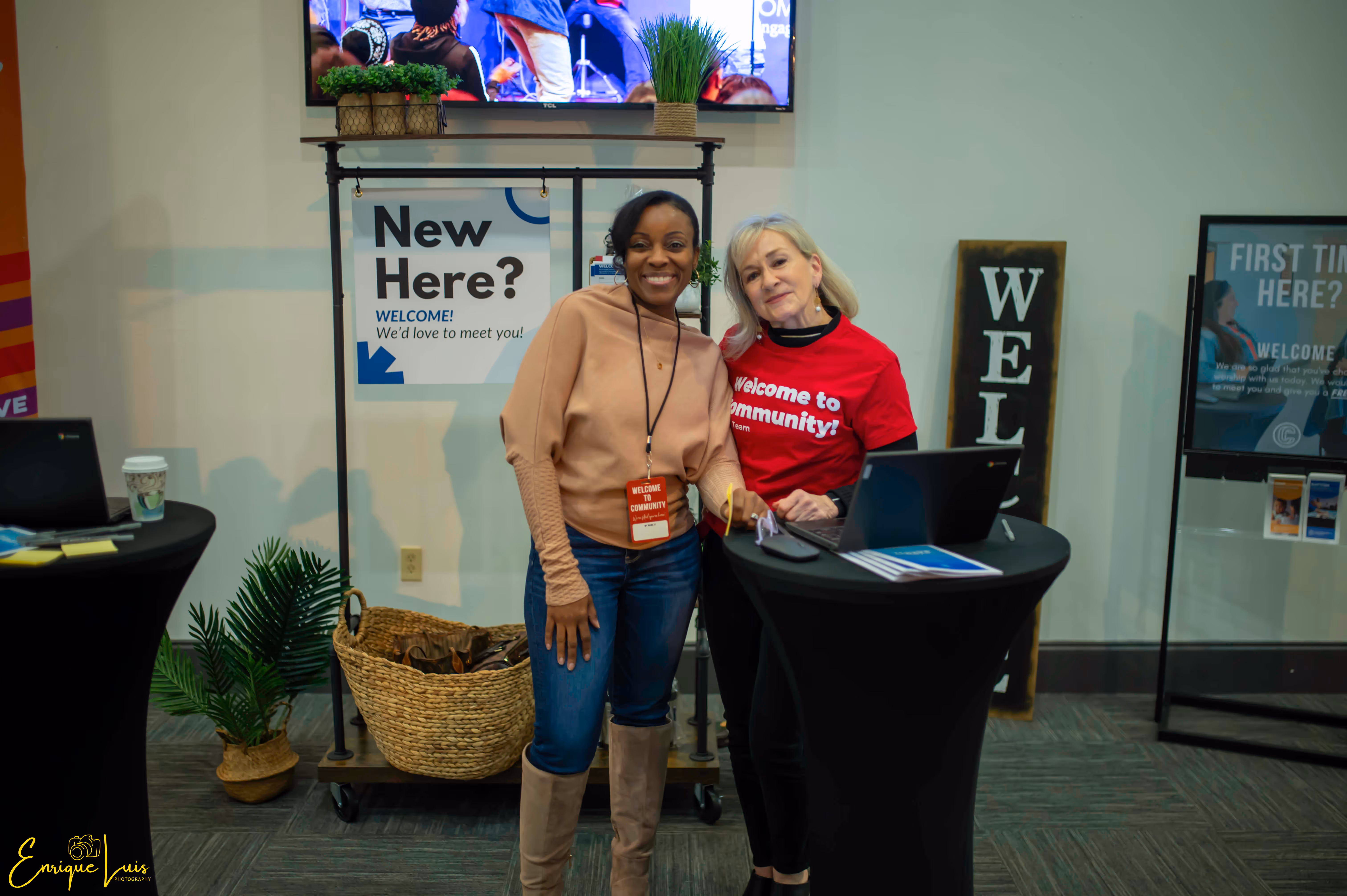 Two women smiling at a welcome desk in a community center, one wearing a red 'Welcome to community!' t-shirt, with signs welcoming new visitors in the background.