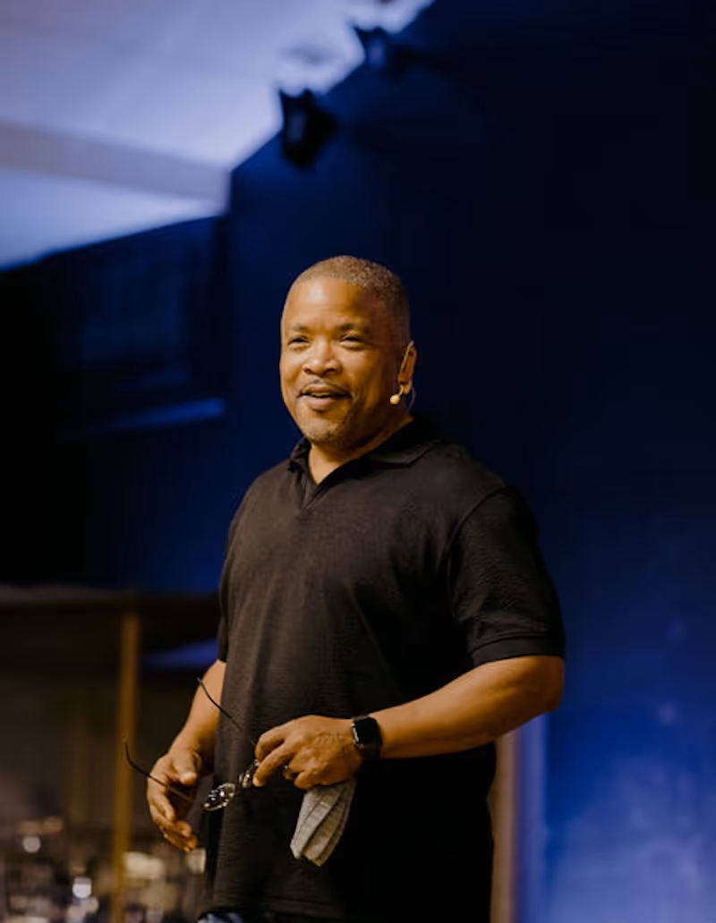 Man wearing a black polo shirt and headset microphone speaking on stage, holding glasses and a cloth.