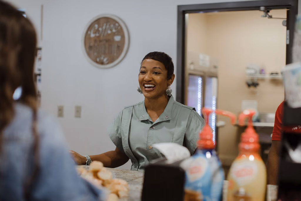 A smiling woman wearing a light green buttoned shirt stands behind a counter, engaging warmly with a person whose back is to the camera, in a cozy indoor setting.