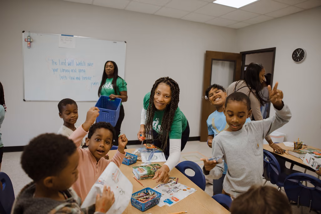 A group of children and two adults engage in an arts and crafts activity around tables in a classroom. One adult wearing a green shirt leans forward smiling, helping the children with their projects. The children hold up drawings and colored pencils, looking happy and excited. A whiteboard and a clock are visible on the walls behind them.