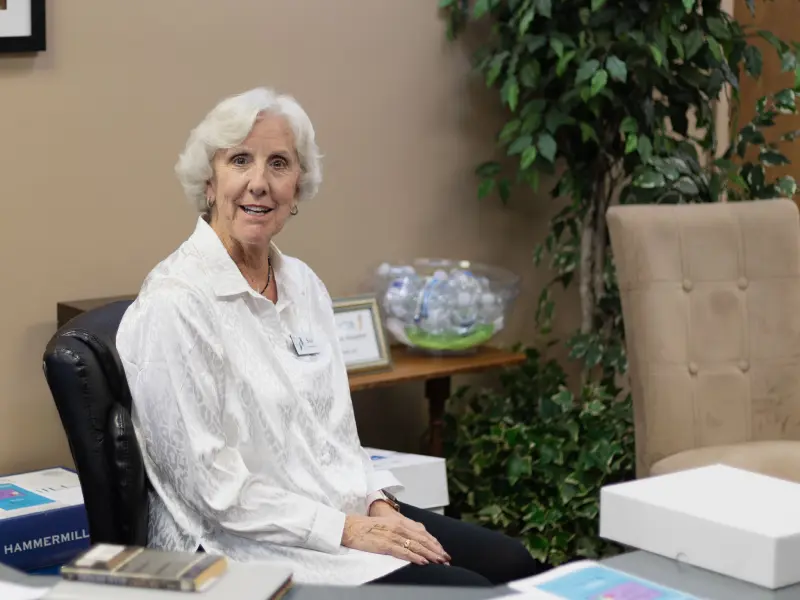 Woman with white hair sitting on a black chair in an office setting, smiling at the camera.