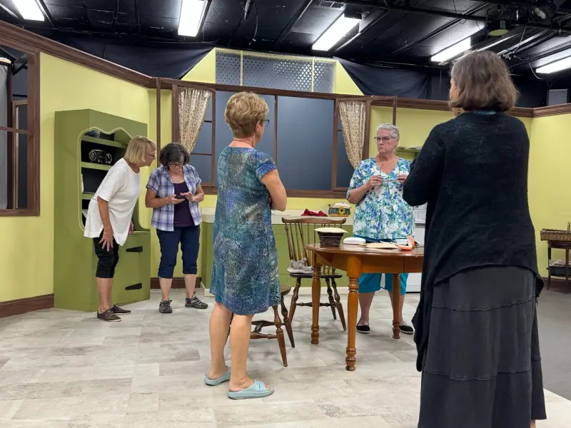 Five women standing and talking in a room with light yellow walls, a wooden table, and chairs.