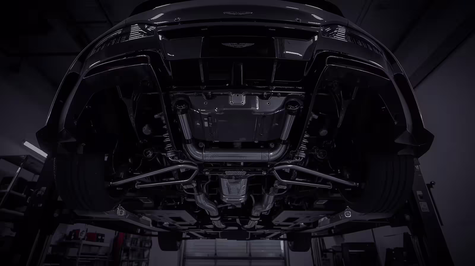 Undercarriage of an Aston Martin on a lift, showing suspension and exhaust layout in black and chrome finish
