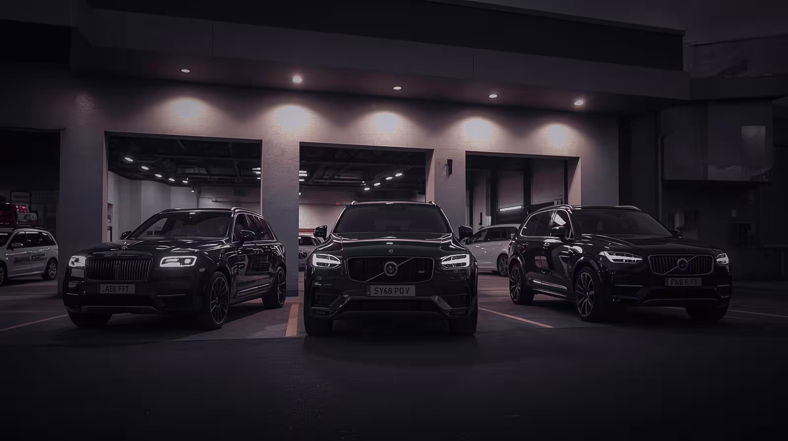 Volvo and Rolls-Royce SUVs parked outside a modern service bay at dusk, headlights illuminated