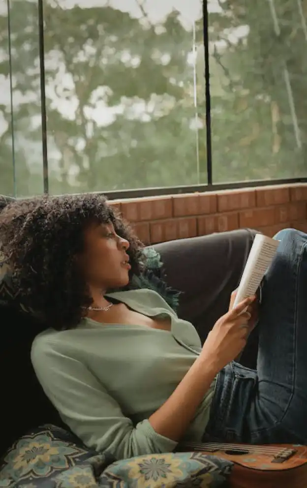 A woman relaxing on a couch while reading a book, representing self-care within the health and wellness industry.