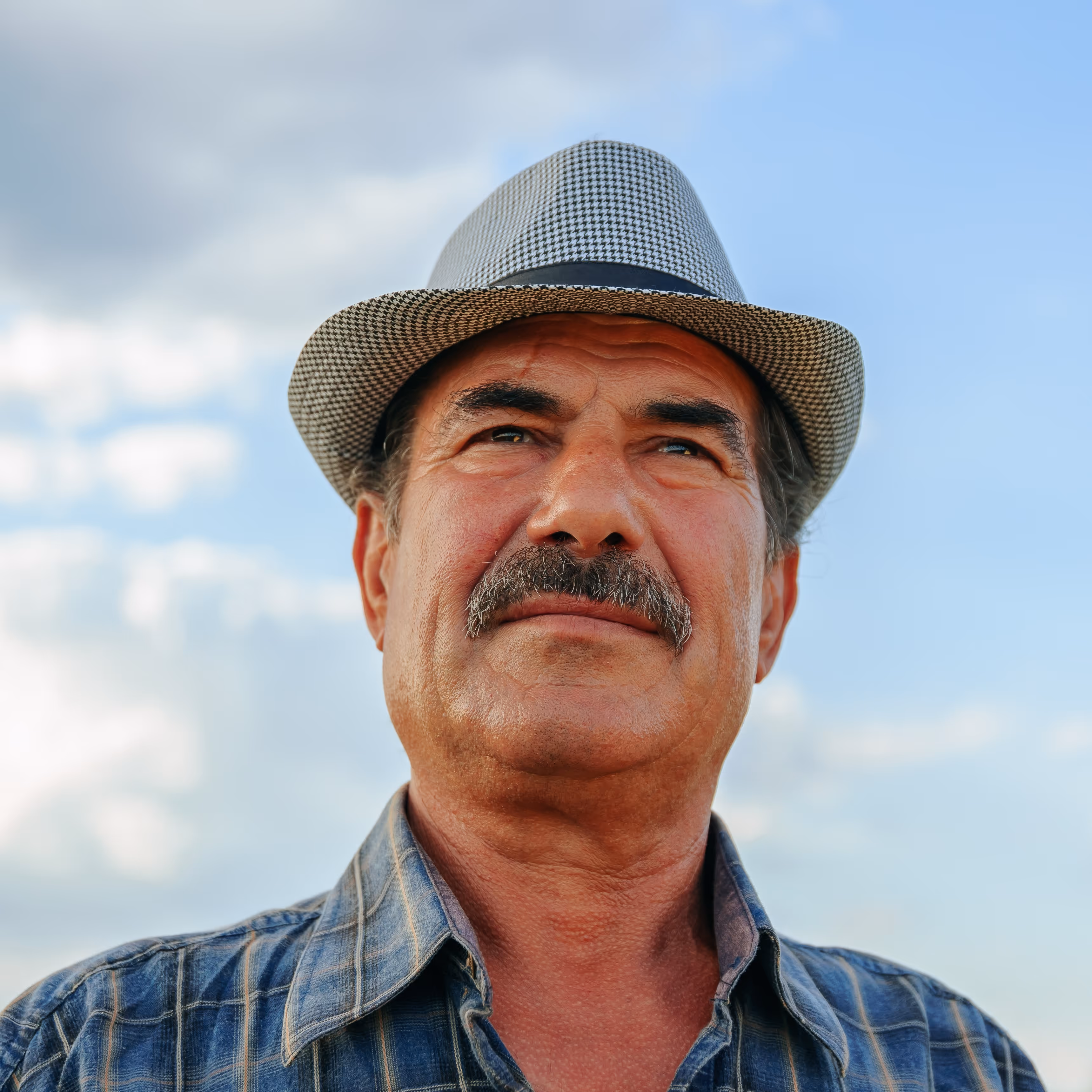 Older man with mustache wearing checkered hat against blue sky