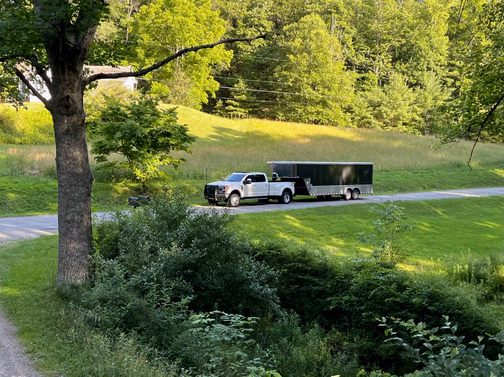 A white pickup truck pulling a long enclosed black trailer drives along a narrow rural road, surrounded by lush green grass and trees.