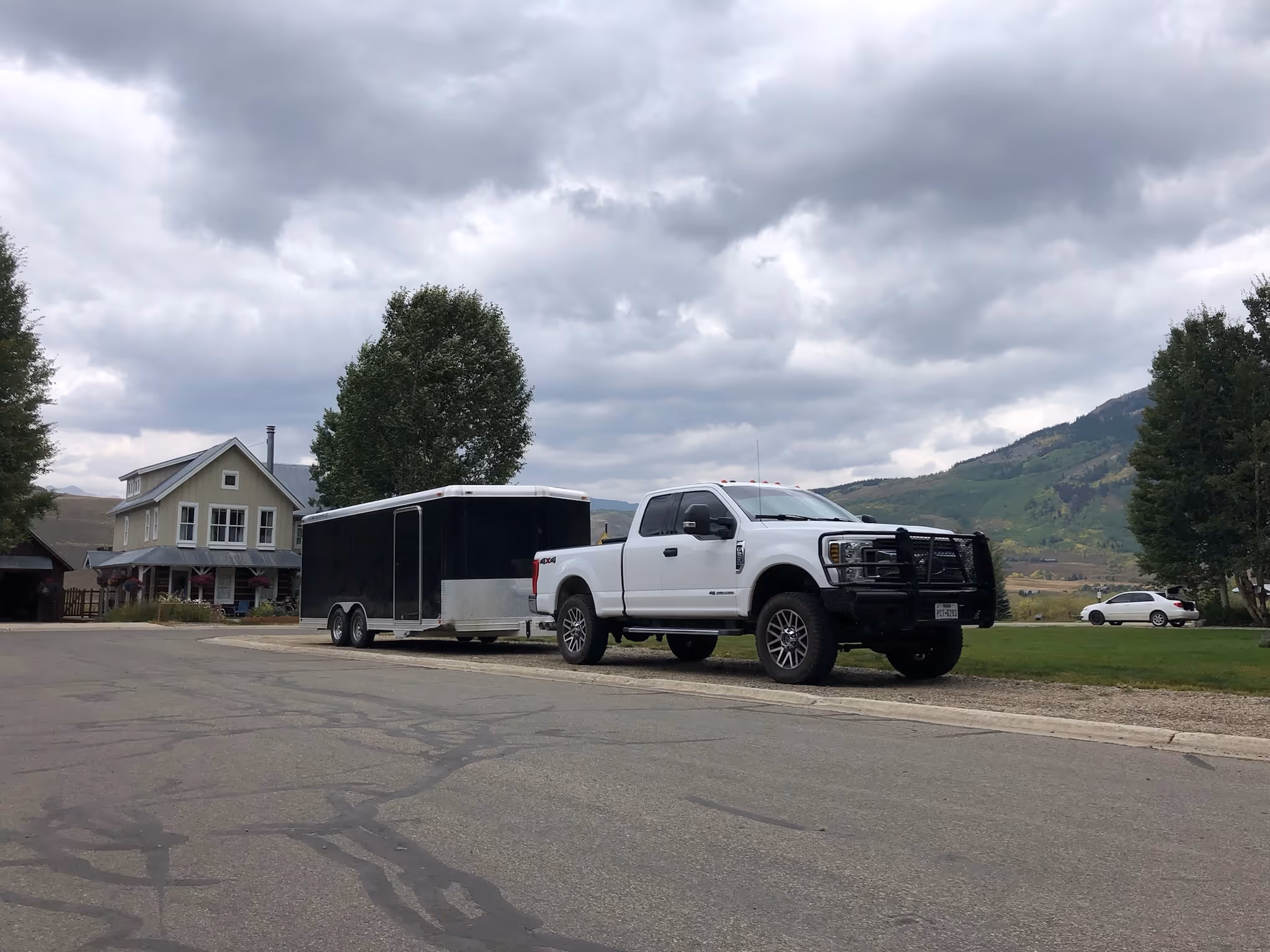 A white pickup truck with a black enclosed trailer is parked on the side of a quiet road in a rural town, with a farmhouse and mountains in the background under a cloudy sky.