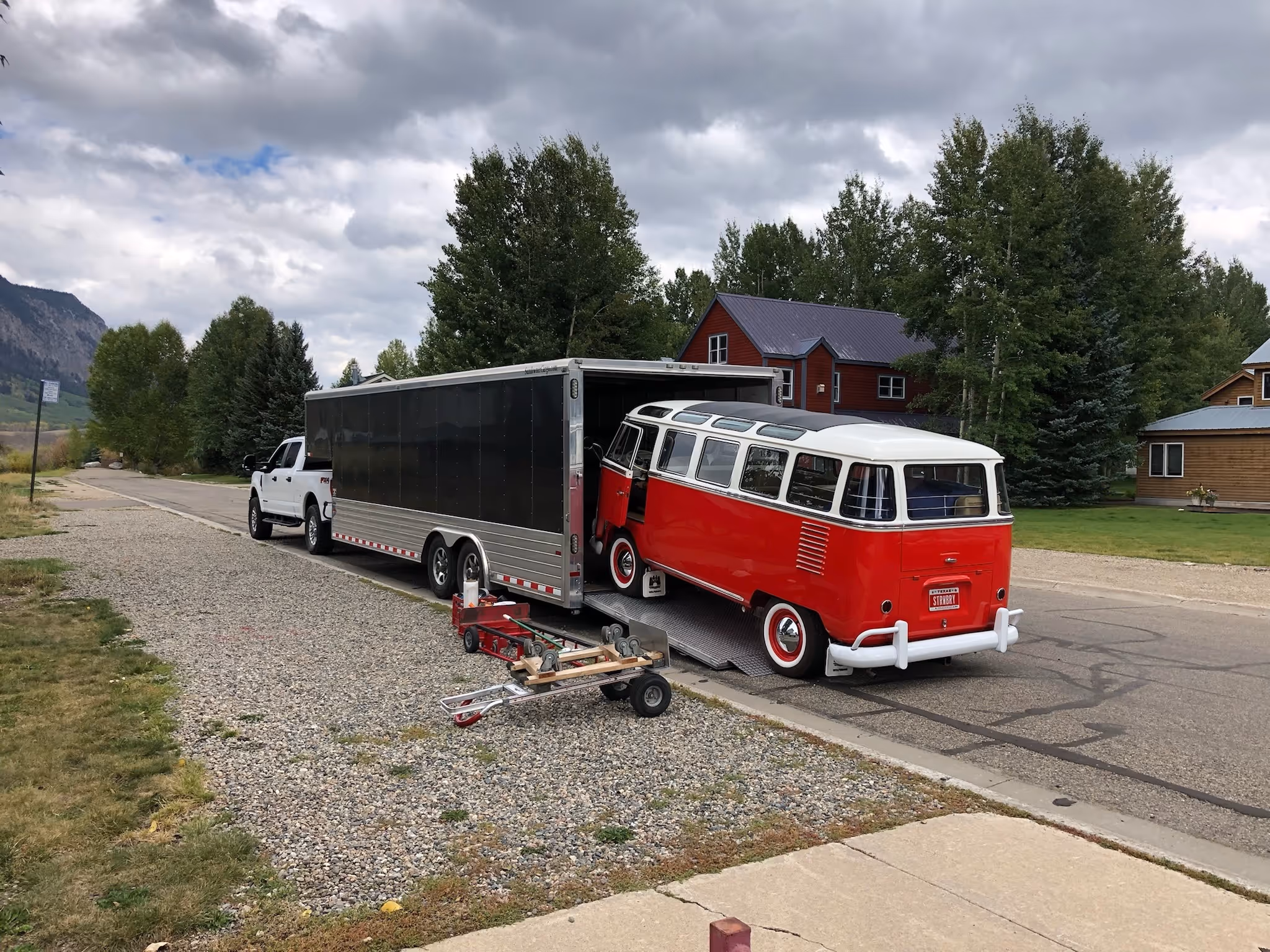 A red and white vintage Volkswagen bus is being loaded onto an enclosed trailer hitched to a white truck, parked on a residential street with trees and houses in the background.