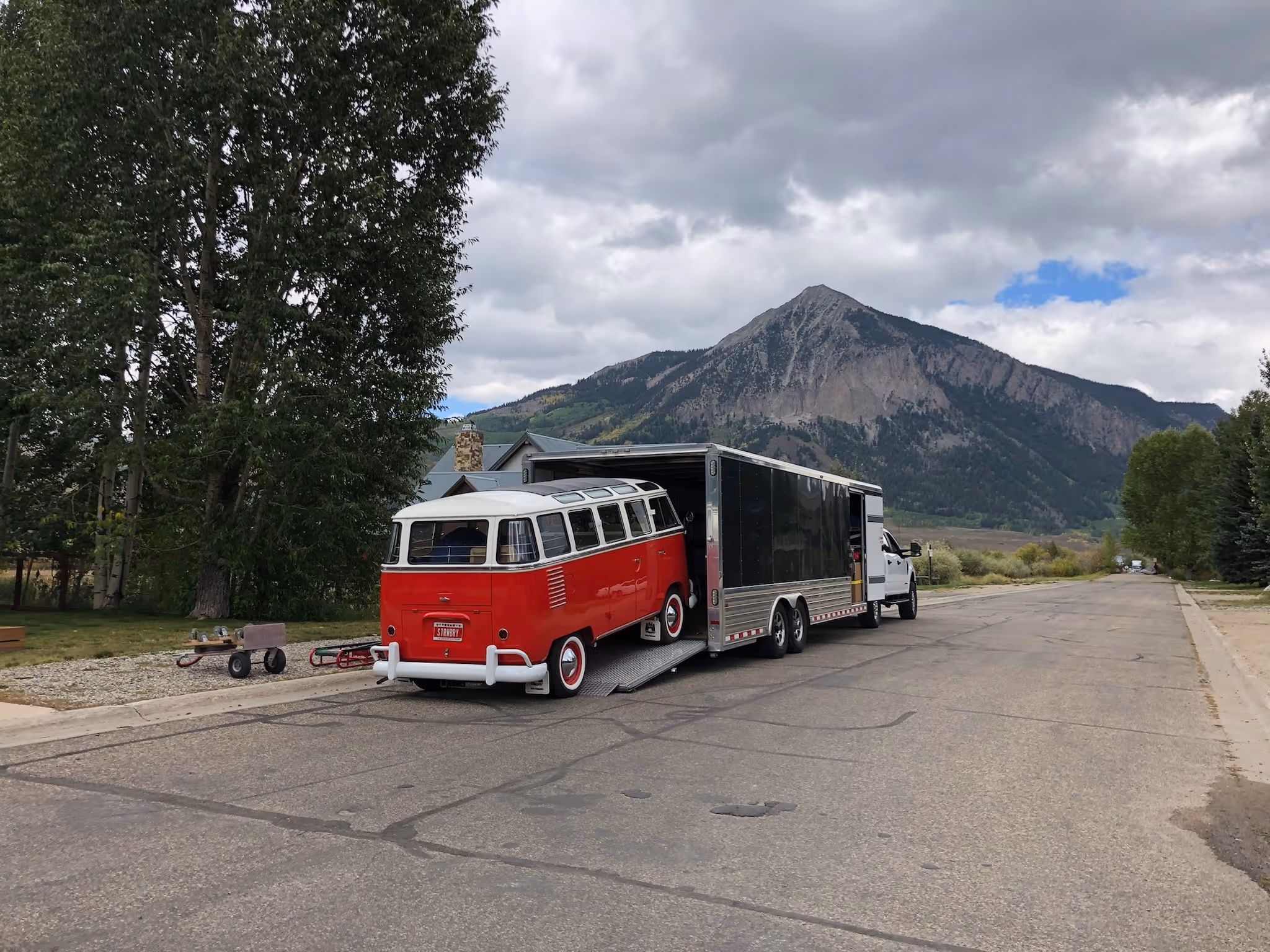 A red and white vintage Volkswagen bus is being loaded into an enclosed trailer attached to a white truck, parked on a quiet street with tall trees and a mountain in the background under a cloudy sky.