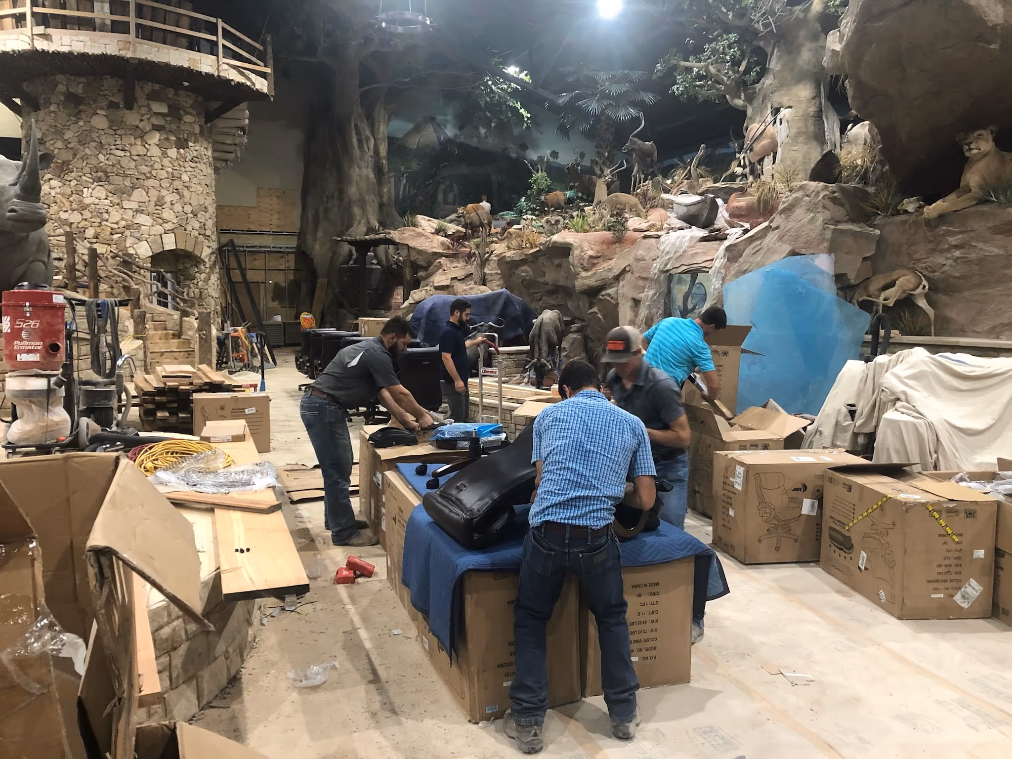 Several workers unpack and set up equipment inside a large indoor space decorated like a natural habitat, with artificial rocks, trees, and mounted animal figures. Cardboard boxes and tools are spread around the work area.