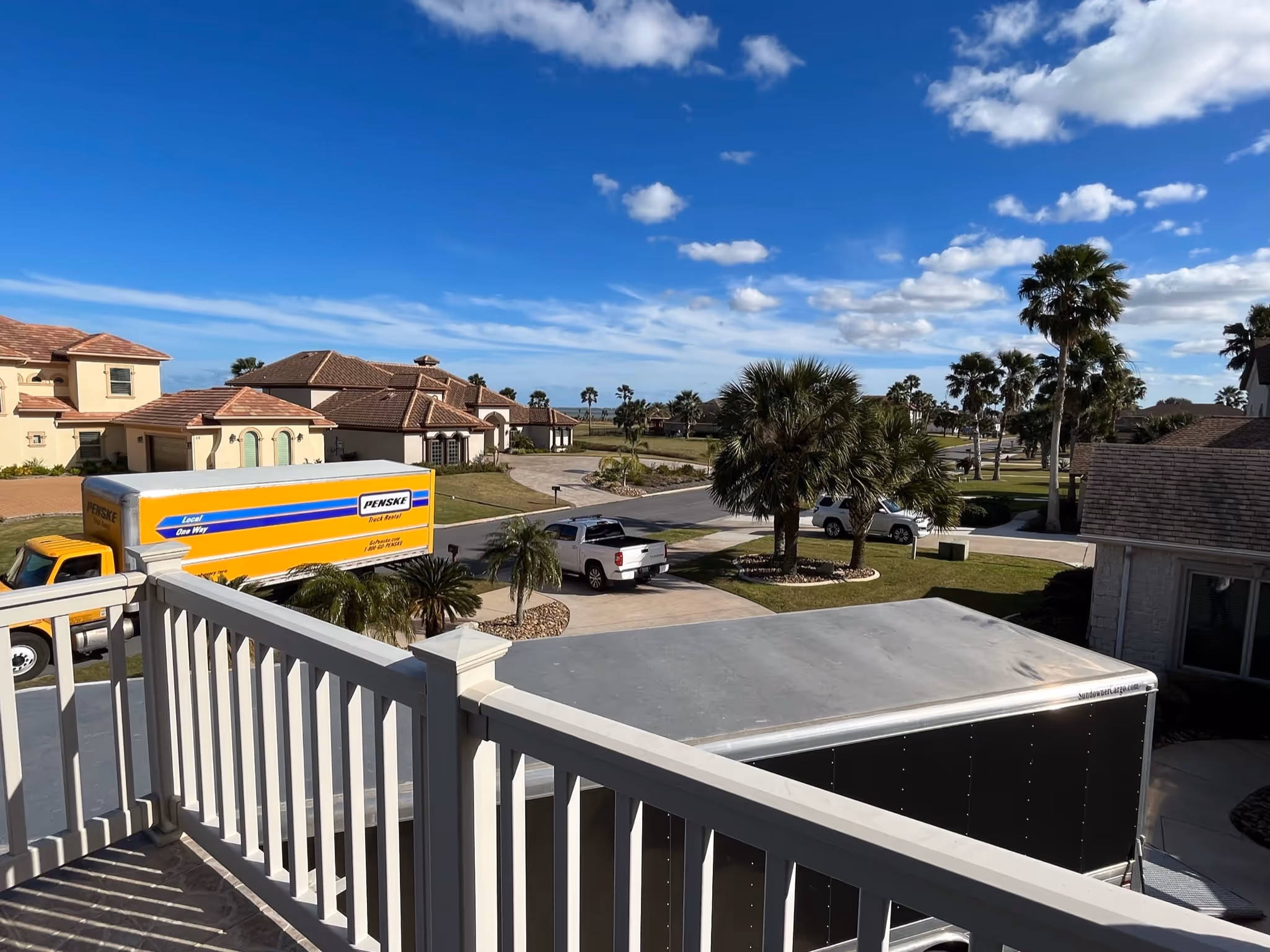 Neighborhood street view with moving trucks, pickup trucks, palm trees, and houses under blue sky.
