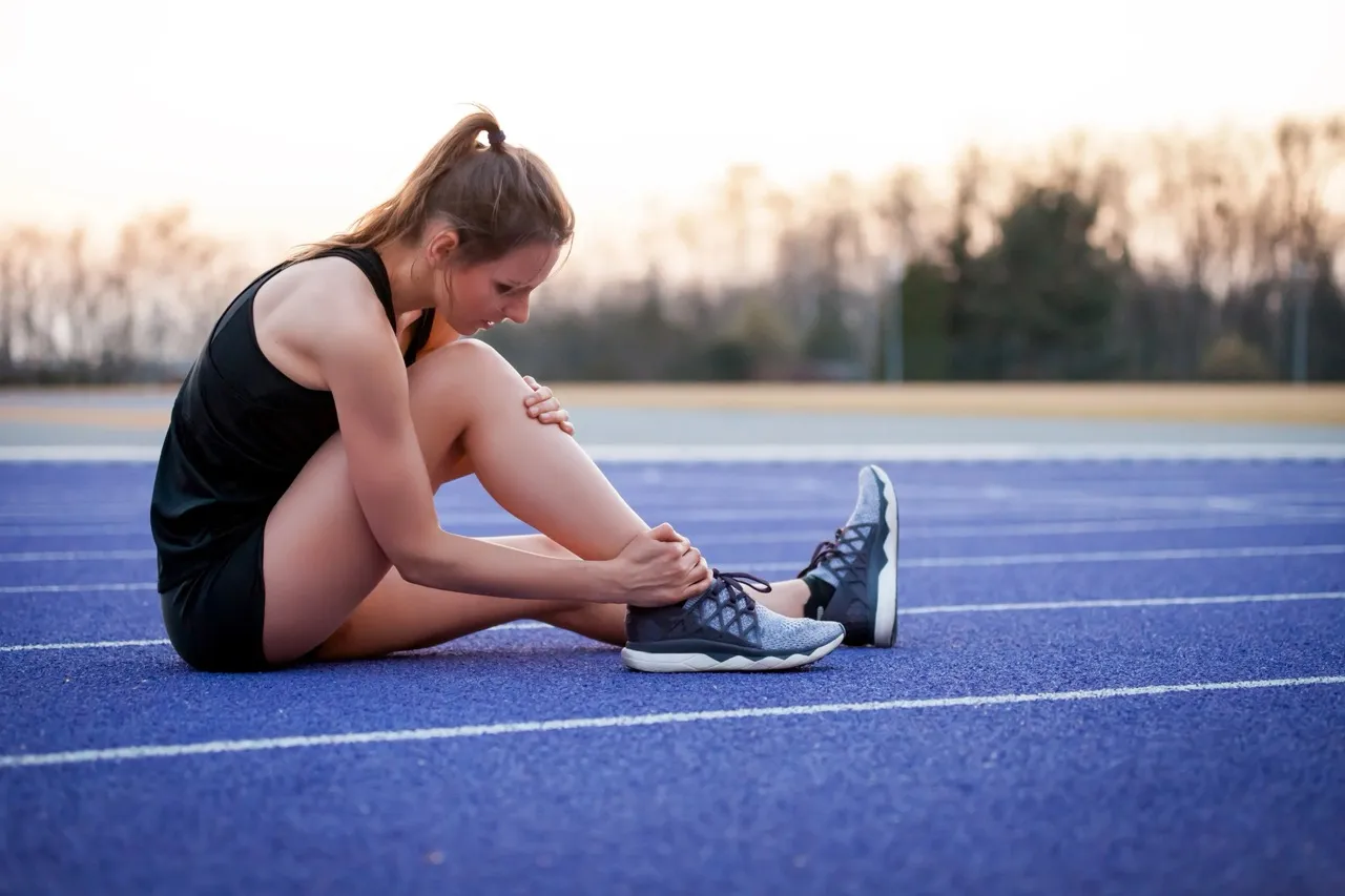 Female athlete sitting on a blue running track, holding ankle after a sports injury, wearing black athletic outfit and running shoes.